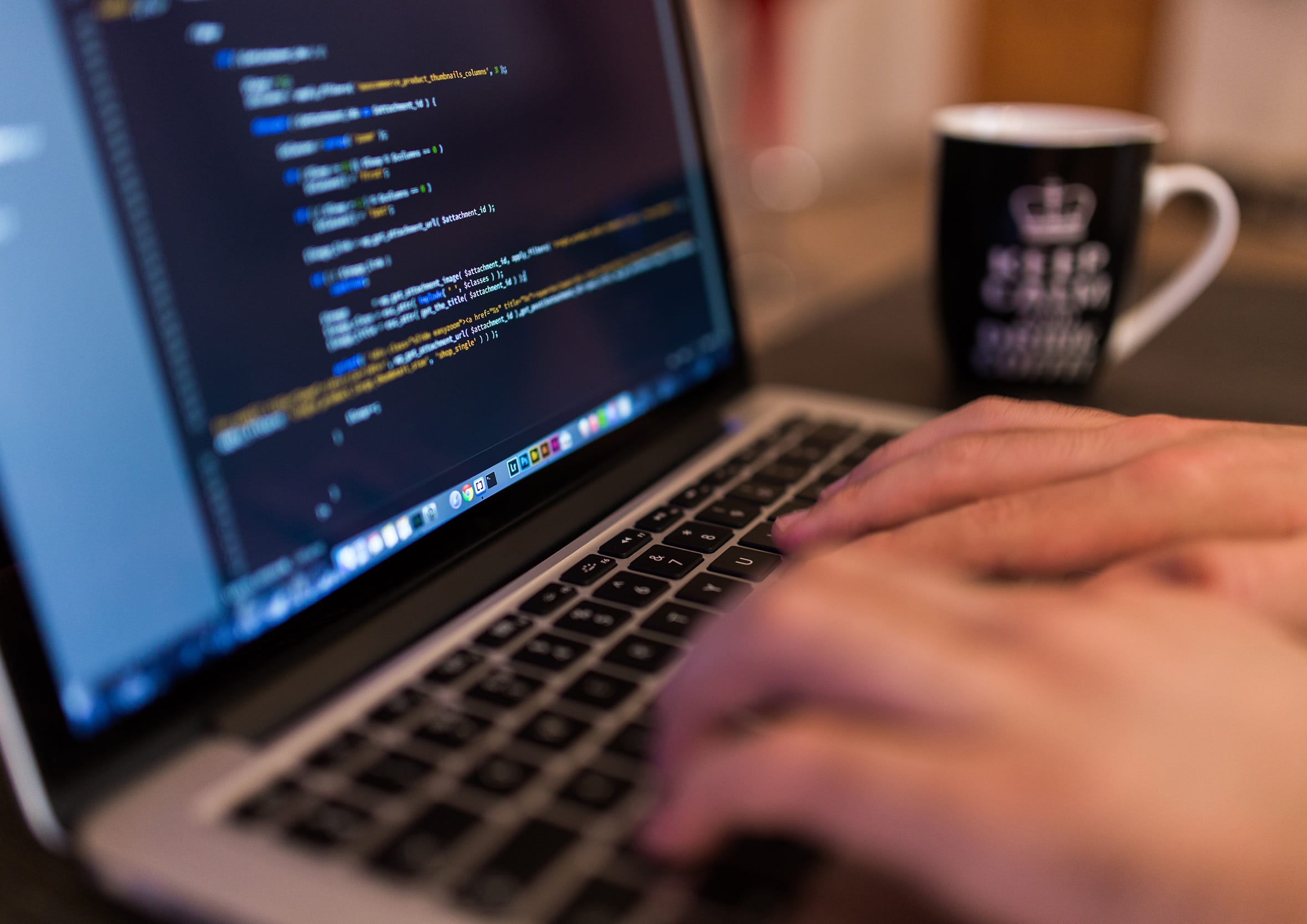 Close-up photograph, hands typing on a laptop keyboard displaying lines of colourful code editor script beside a dark coffee mug, illustrating hands-on software engineering and technical build execution.
