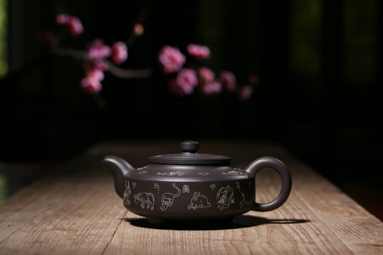 Dark clay teapot with detailed Chinese zodiac line engravings resting on a rustic wooden table with soft blossom bokeh, highlighting craftsmanship and object design.