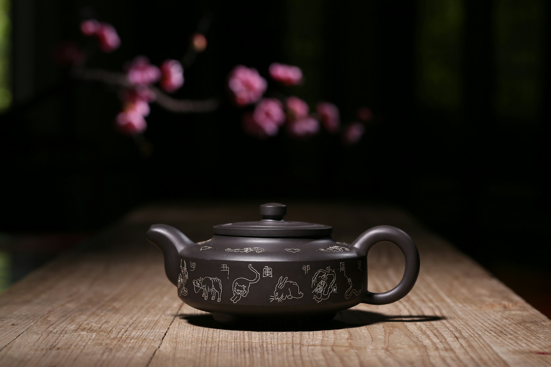 Dark clay teapot with detailed Chinese zodiac line engravings resting on a rustic wooden table with soft blossom bokeh, highlighting craftsmanship and object design.