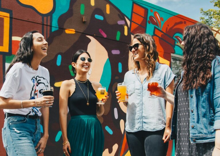 Group of four friends laughing and holding craft beer glasses outside in front of a colourful street mural, capturing social connection and relaxed lifestyle moments.