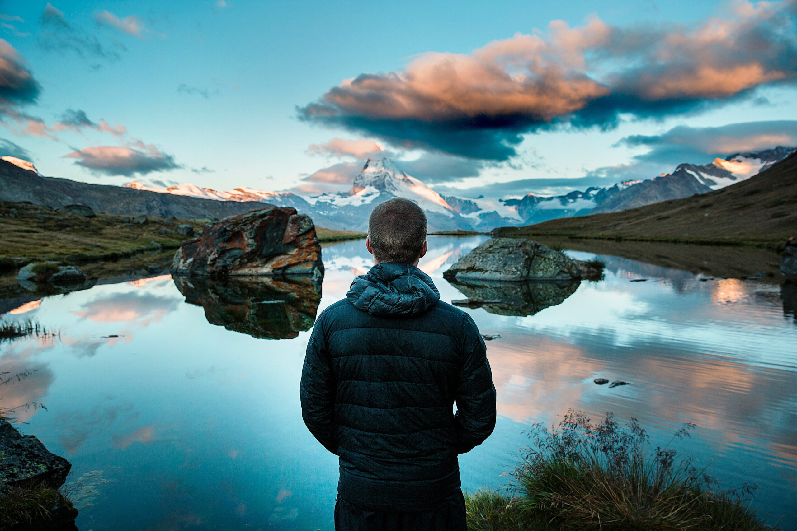 Person seen from behind in a padded jacket looking out at a calm alpine lake reflecting a snow-capped mountain peak and scattered clouds.