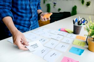 Close-up of a designer organising paper index cards with handwritten labels on a desk during a UX card-sorting exercise, demonstrating information architecture synthesis.