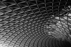 Black and white photograph of a sweeping, geometric lattice roof frame, showing intricate metalwork and modern structural architecture.