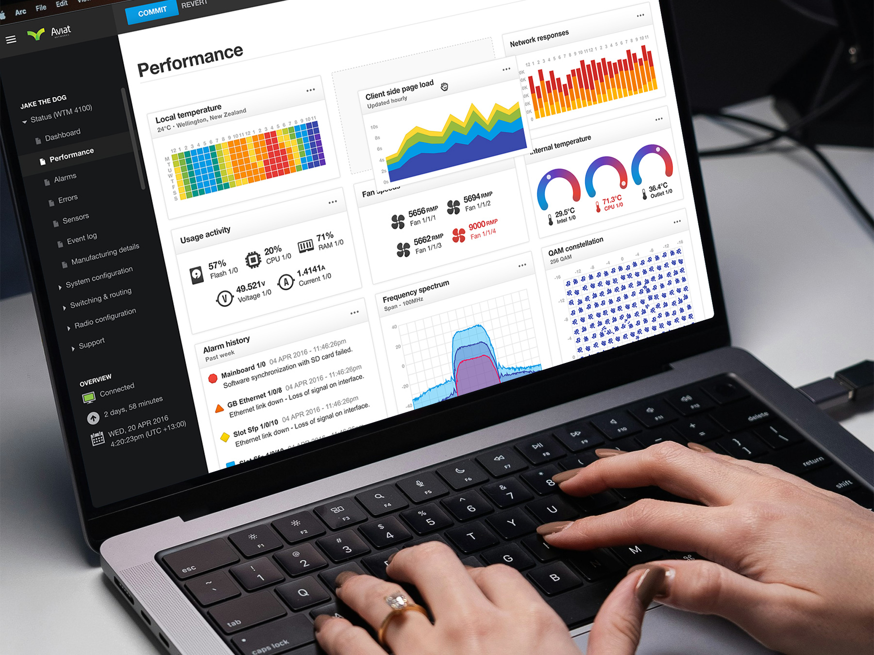 Hands typing on a laptop keyboard displaying the Aviat Networks radio router performance dashboard UI with metrics for usage, temperature, frequency spectrum, and system alarms.