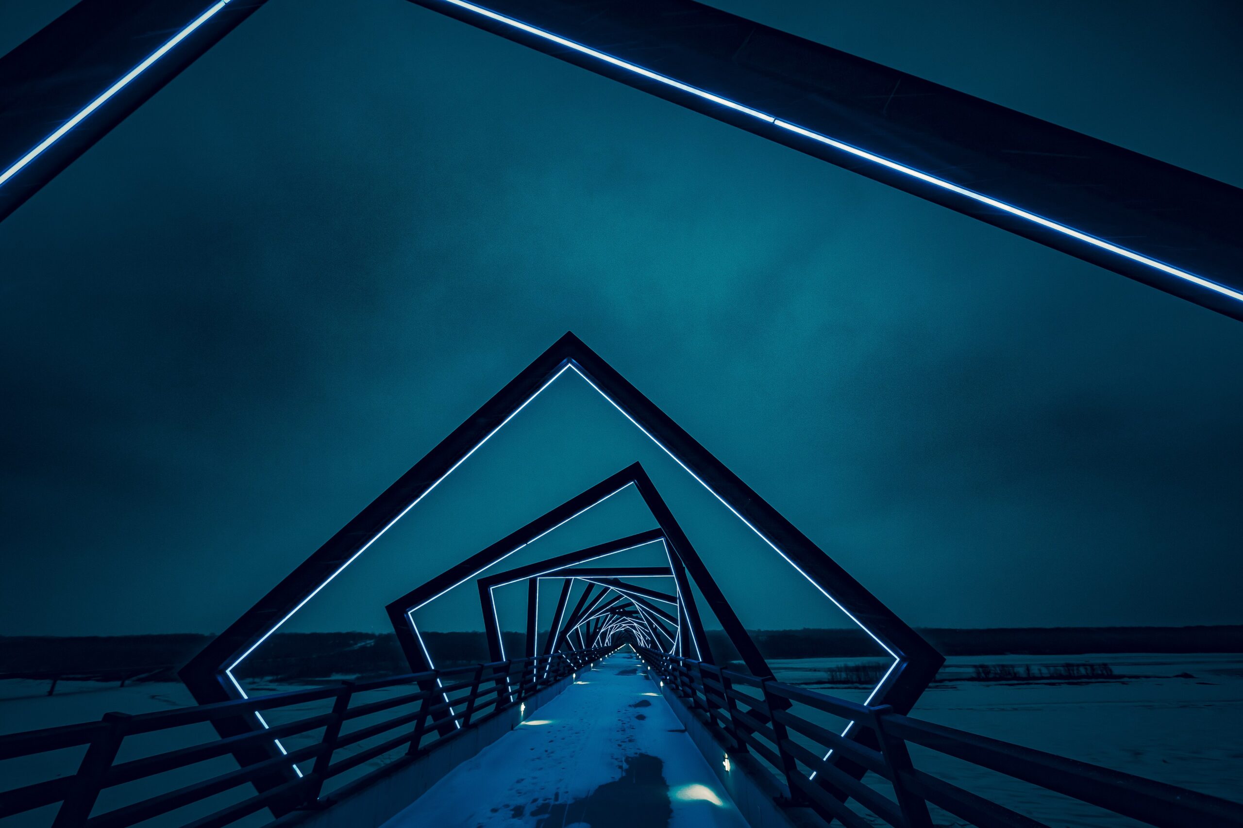 Night perspective looking through the glowing blue geometric steel arches of the High Trestle Trail Bridge, showcasing modern architectural lighting and linear perspective.