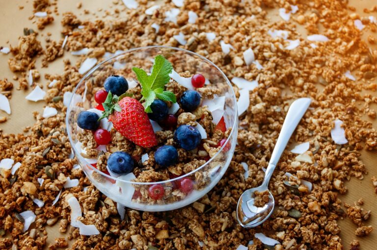 Clear bowl filled with yogurt, crunchy muesli, blueberries, and a fresh strawberry set against a scattered granola background, highlighting healthy breakfast concepts.