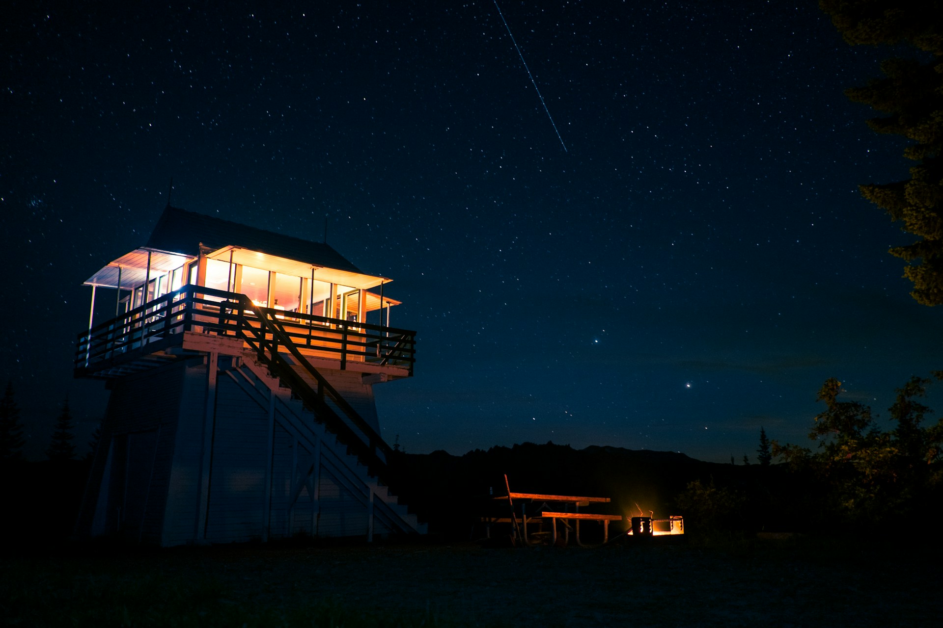 Illuminated wooden fire lookout tower at night under a star-filled sky with a glowing campfire below, illustrating outdoor solitude and astrophotography.