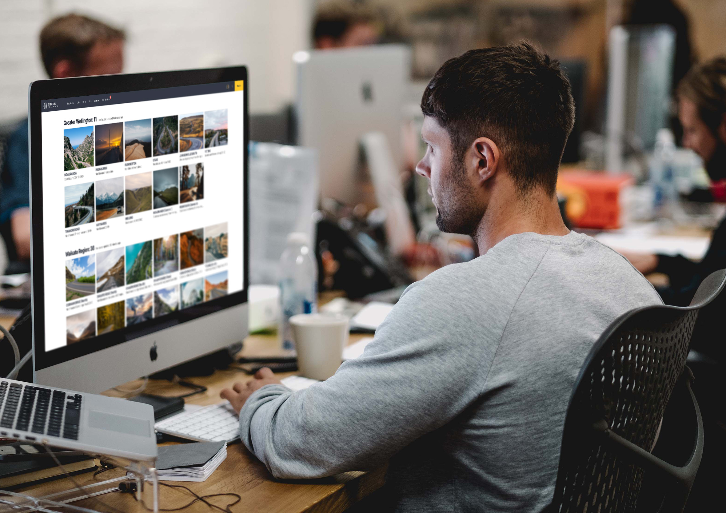 Side view mockup of a designer working at a desk setup with an iMac displaying the dispatch dashboard CCTV camera gallery.