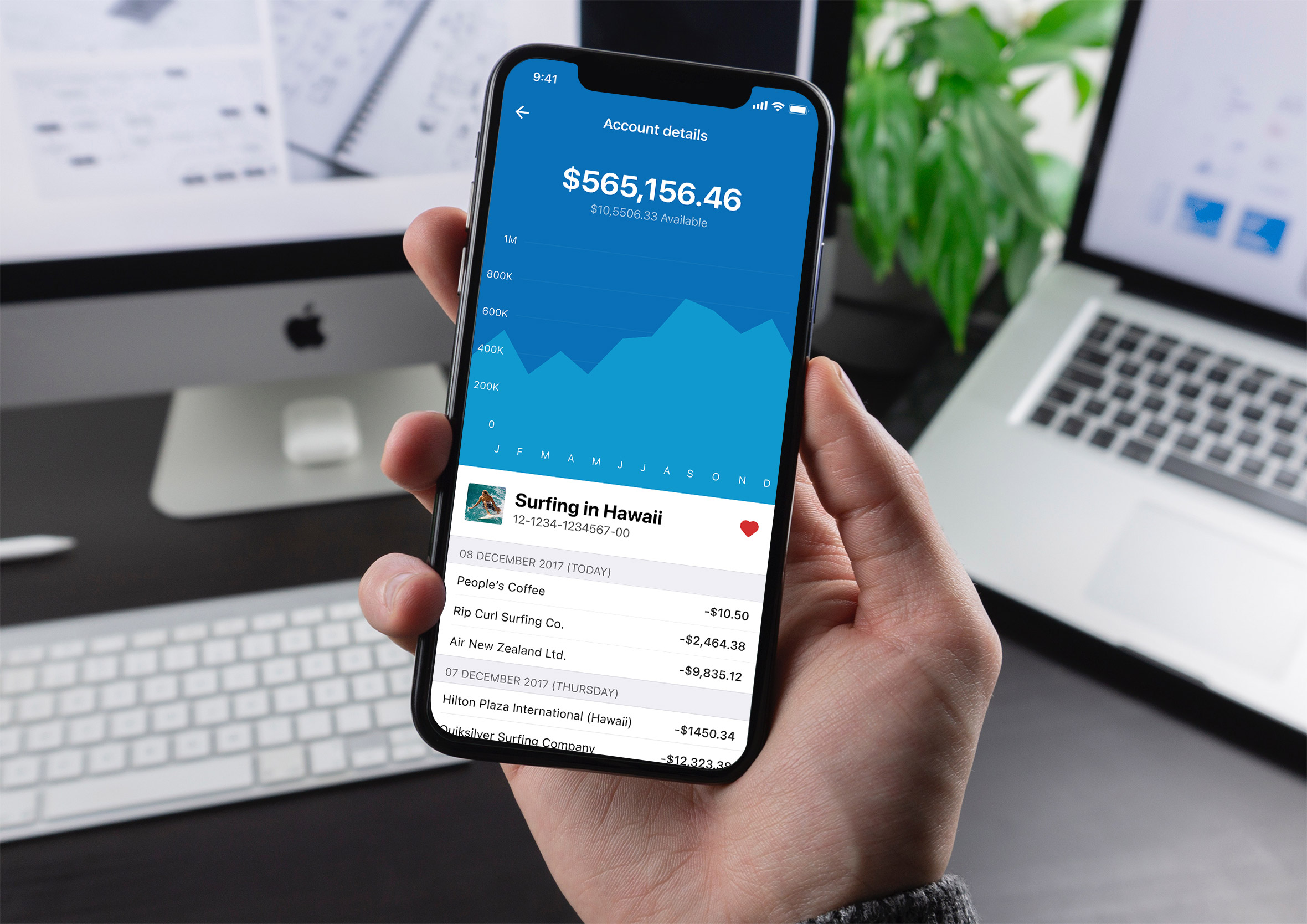 Hand holding an iPhone displaying a core banking app's account details screen showing total balance, savings graph, and recent transaction history at a desk.