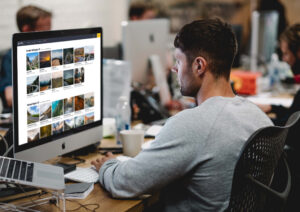 A designer seated at a wooden desk working on a desktop iMac in a busy studio office environment.