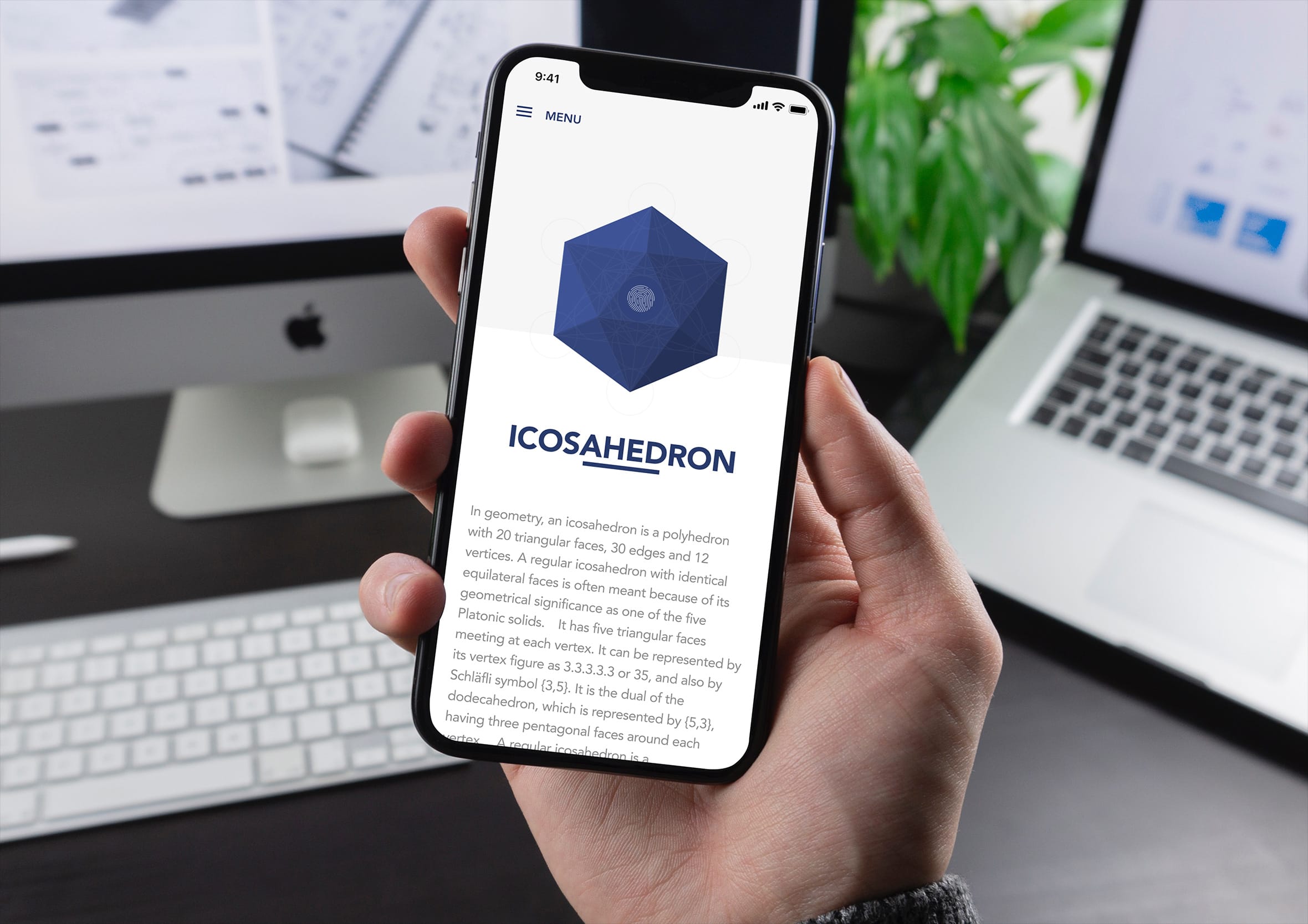 Hand holding a smartphone displaying the blue icosahedron shape detail page on a dark desk with desktop screens in the background.