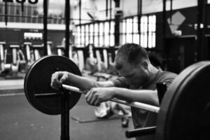 Black and white photo of a man leaning exhausted against a loaded barbell in a gym, highlighting physical training and discipline.