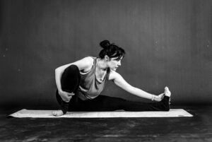 Black and white shot of a woman performing a seated leg stretch on a yoga mat, illustrating mobility and physical wellness routines.