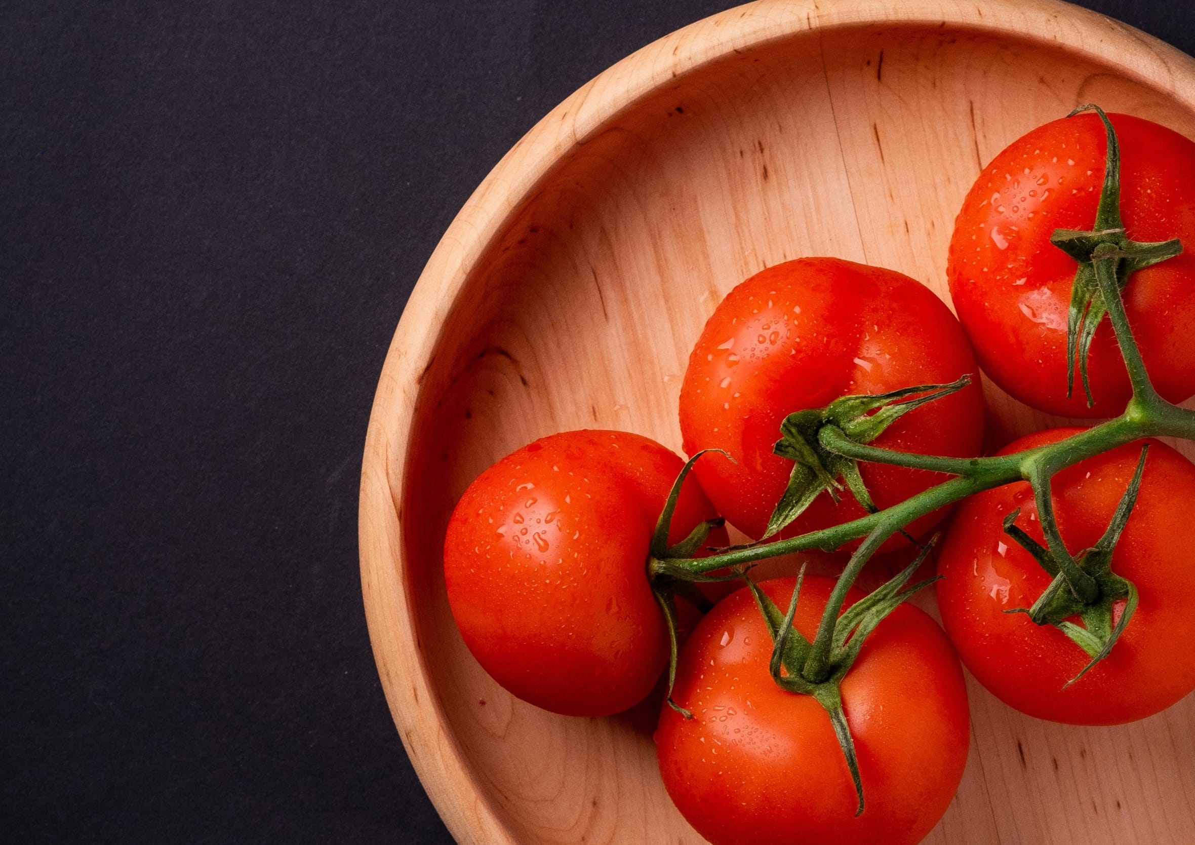 Cluster of fresh red vine tomatoes sitting in a natural wooden bowl on a dark background