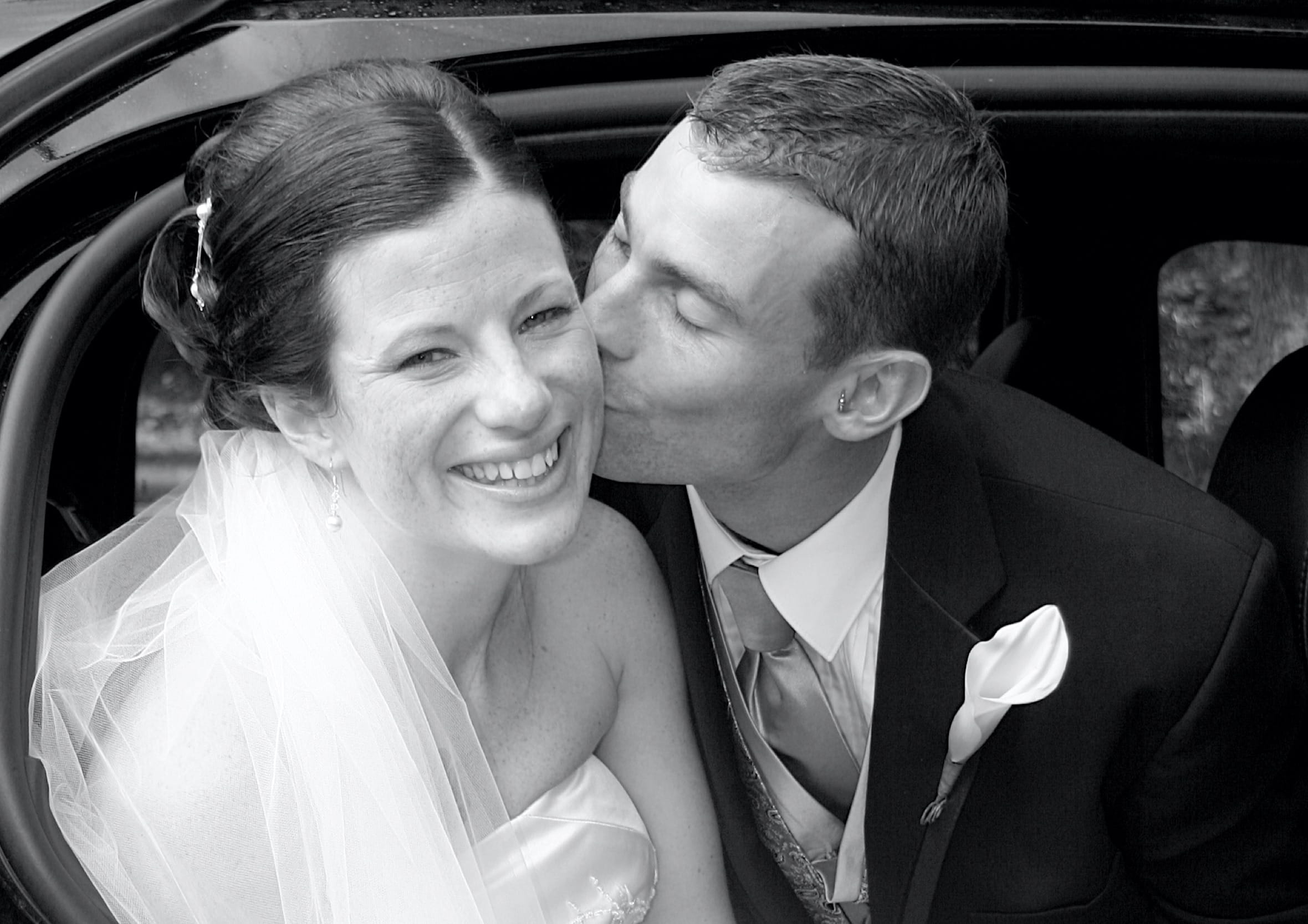 Black and white photo of a groom kissing the bride on her cheek inside a wedding car