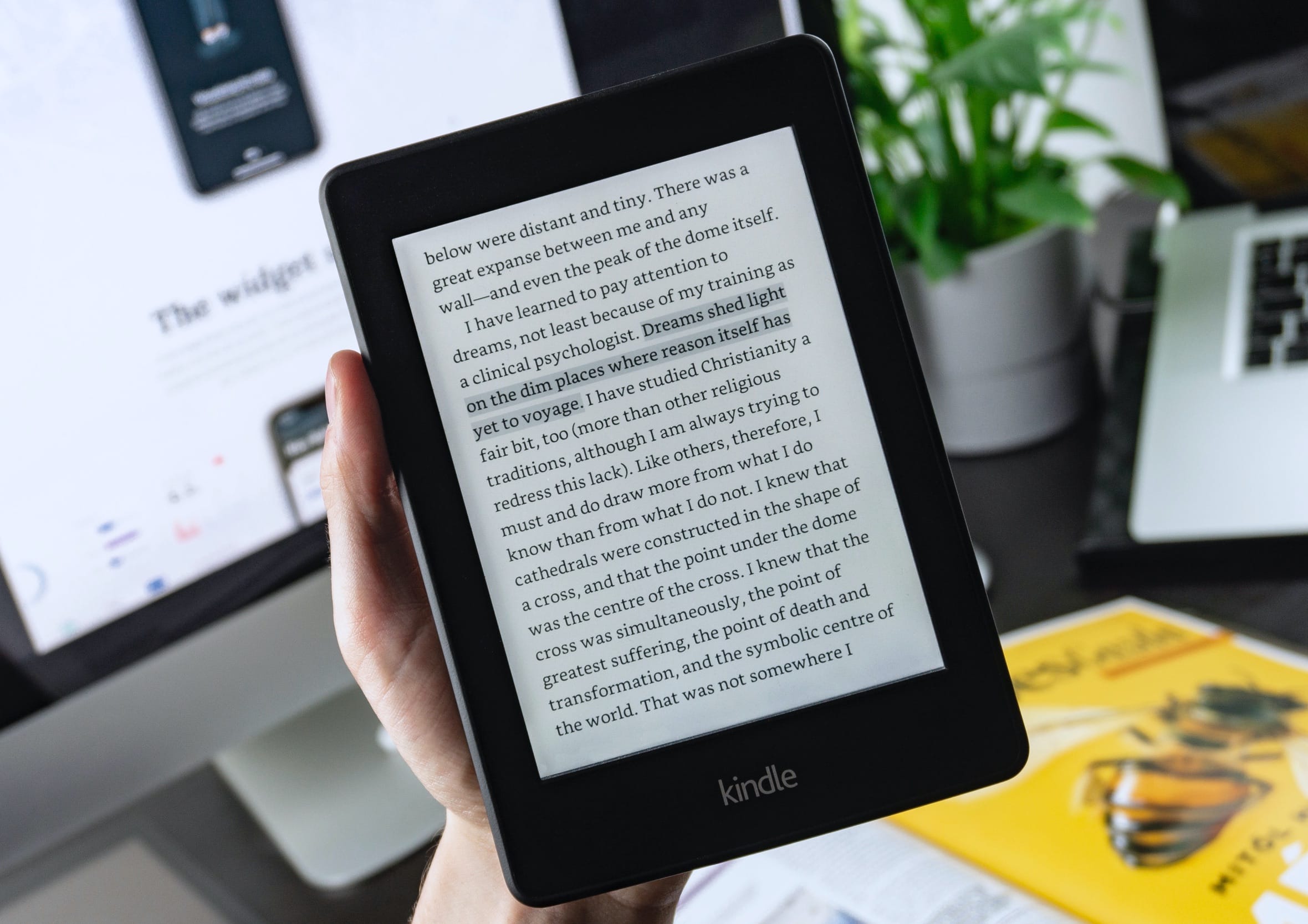 Hand holding an Amazon Kindle e-reader displaying crisp digital body text in front of a modern workspace for portable, distraction-free reading.