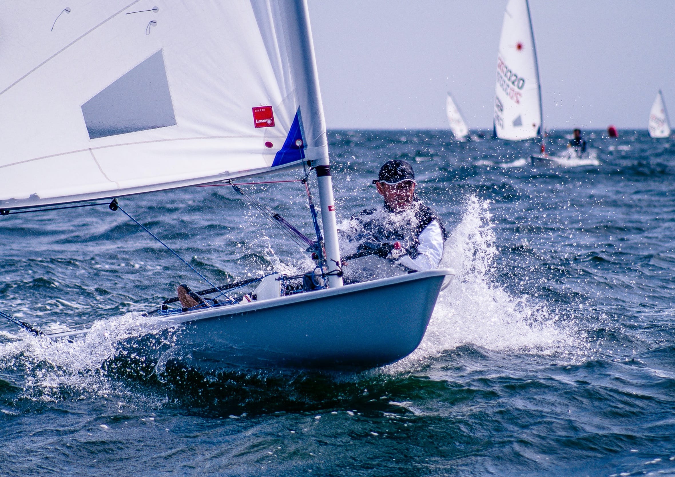 Sailor hiking out on a small Laser sailboat while cutting through splashing ocean waves