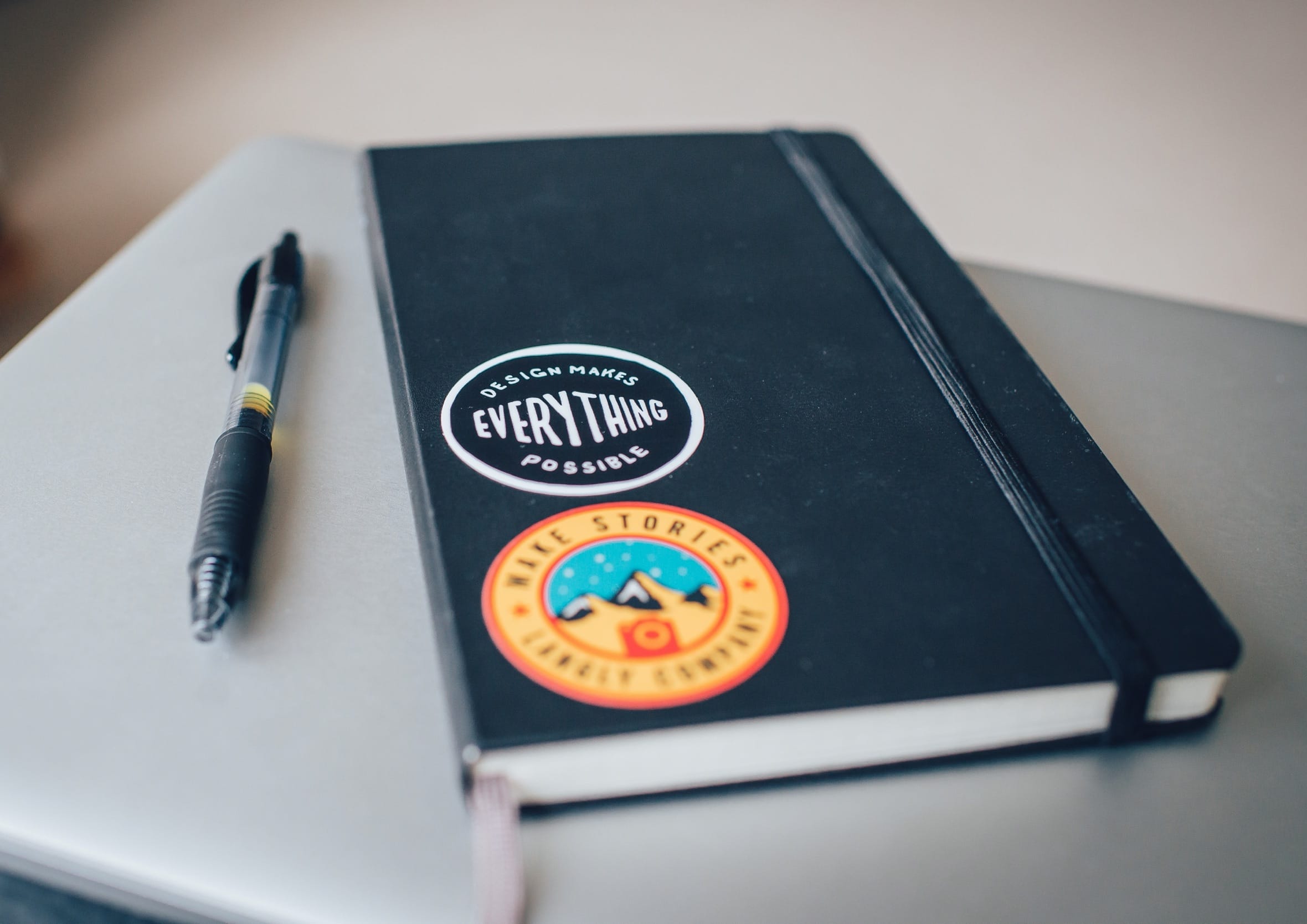Black hardbound Moleskine notebook sitting on a laptop with stickers reading "Design Makes Everything Possible" alongside a black ballpoint pen for analog sketching and idea collection.