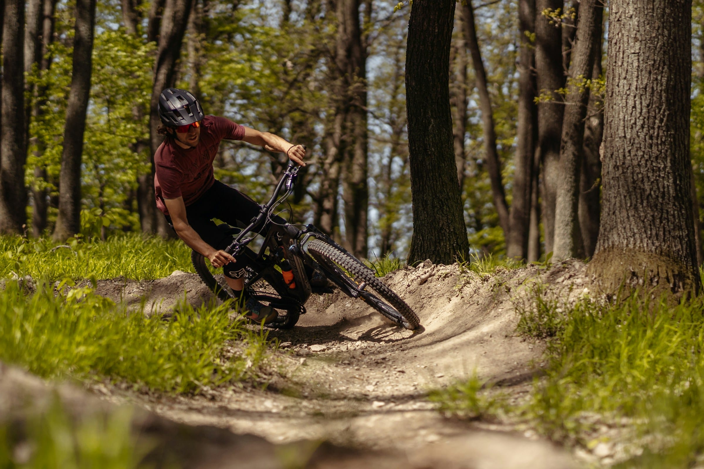 Action photo of a mountain biker catching air over a dirt jump on a wooded trail.