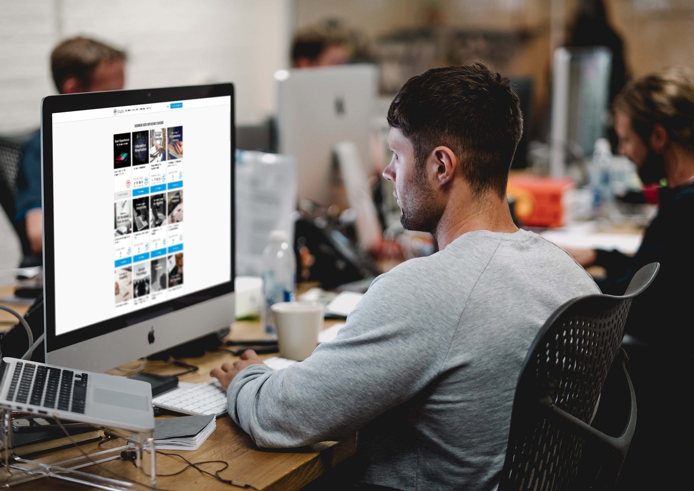 Designer sitting at an office desk working on an iMac monitor displaying an online learning course layout with course cards and UI components for skill development.
