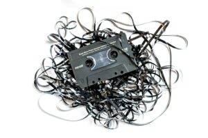 Black cassette tape lying on a white background surrounded by a tangled heap of unspooled magnetic tape, representing analogue media and audio preservation.