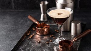 Espresso martini cocktail topped with coffee beans in a martini glass next to copper measuring scoops, highlighting hospitality presentation and craft beverage detail.