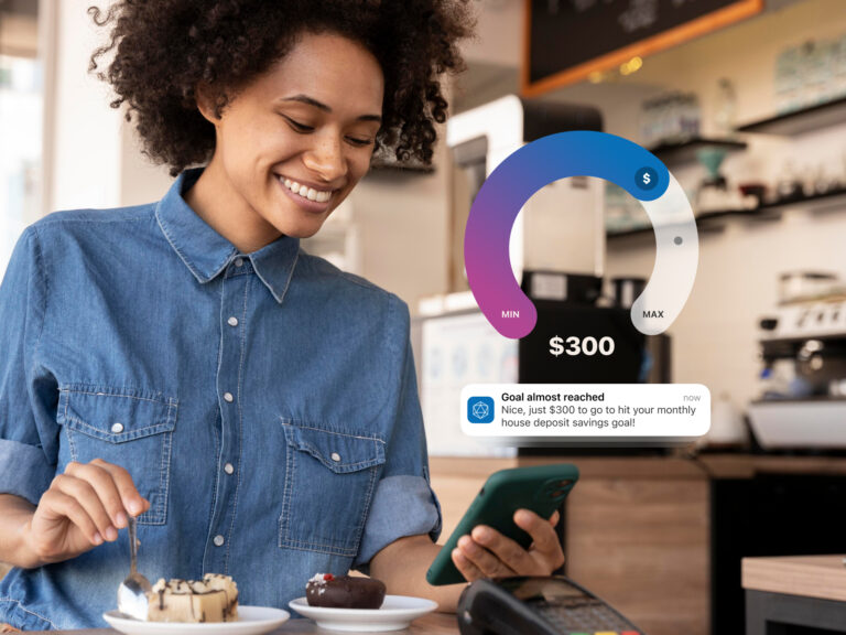 Woman smiling at her phone in a cafe, overlaid with a UI graphic showing a circular savings goal tracker and a notification reading 'Goal almost reached'.