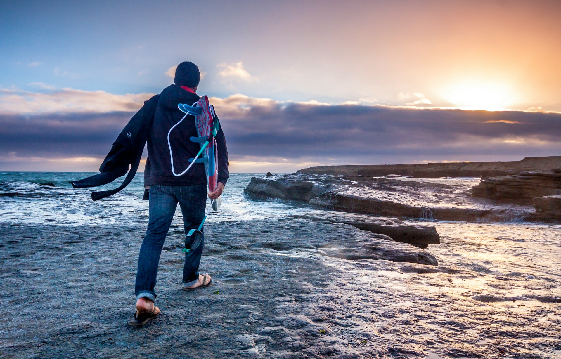 Person in a dark hoodie and jeans carrying a surfboard under their arm while walking across a flat, rocky shore towards a golden sunset.
