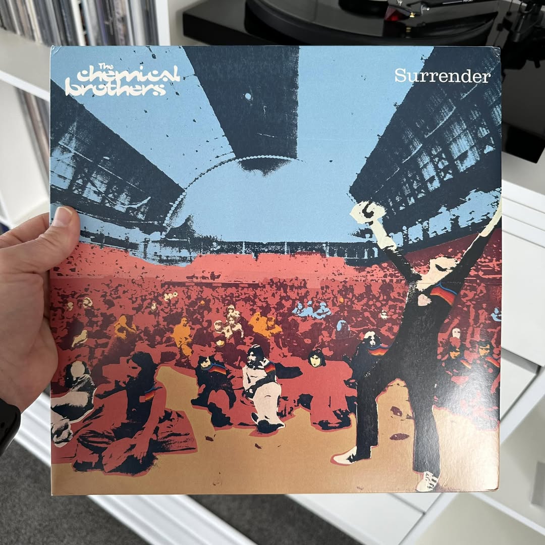 Person holding a vinyl sleeve of The Chemical Brothers' Surrender album featuring stylized screen-printed artwork of a crowd under a light-blue sky with a central figure raising their arms in triumph, providing visual inspiration for graphic contrast and high-energy music aesthetic showcase.