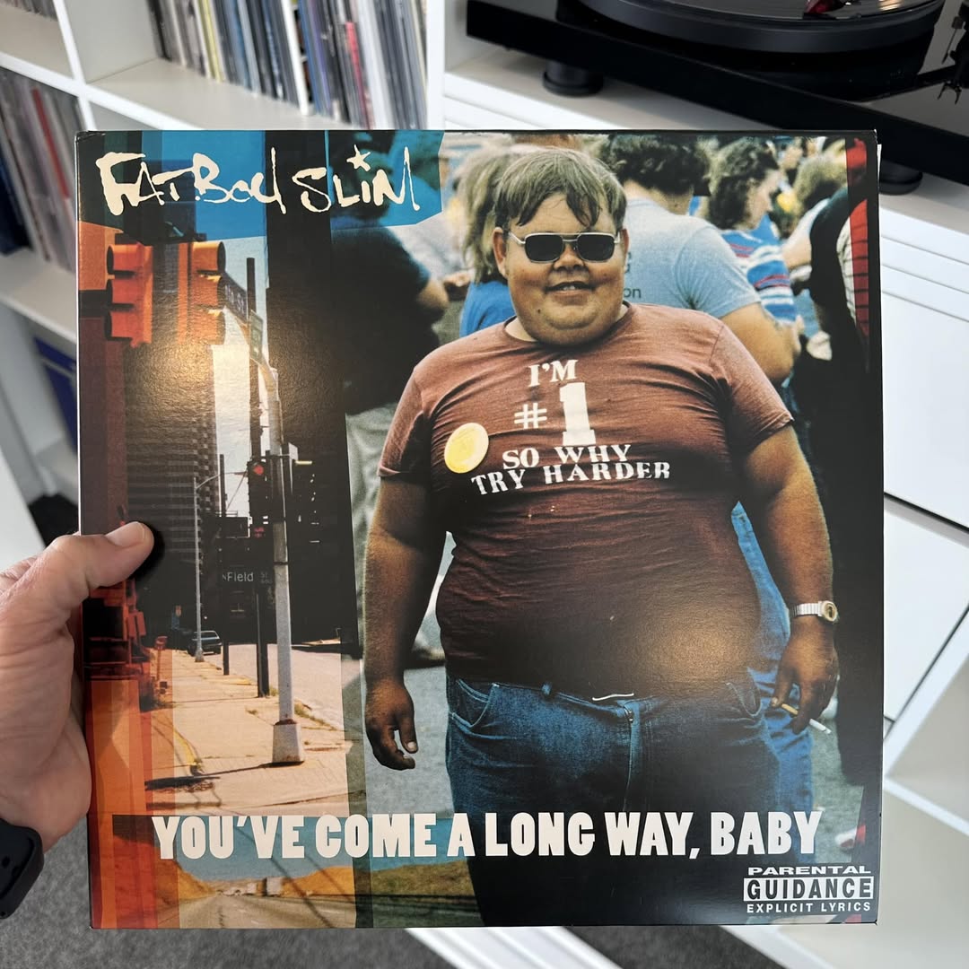 Person holding a vinyl sleeve of Fatboy Slim's You've Come a Long Way, Baby album featuring a large man wearing sunglasses and a T-shirt printed with micro-copy reading "I'm #1 So Why Try Harder", serving as visual inspiration for nostalgic pop-culture aesthetic showcase.