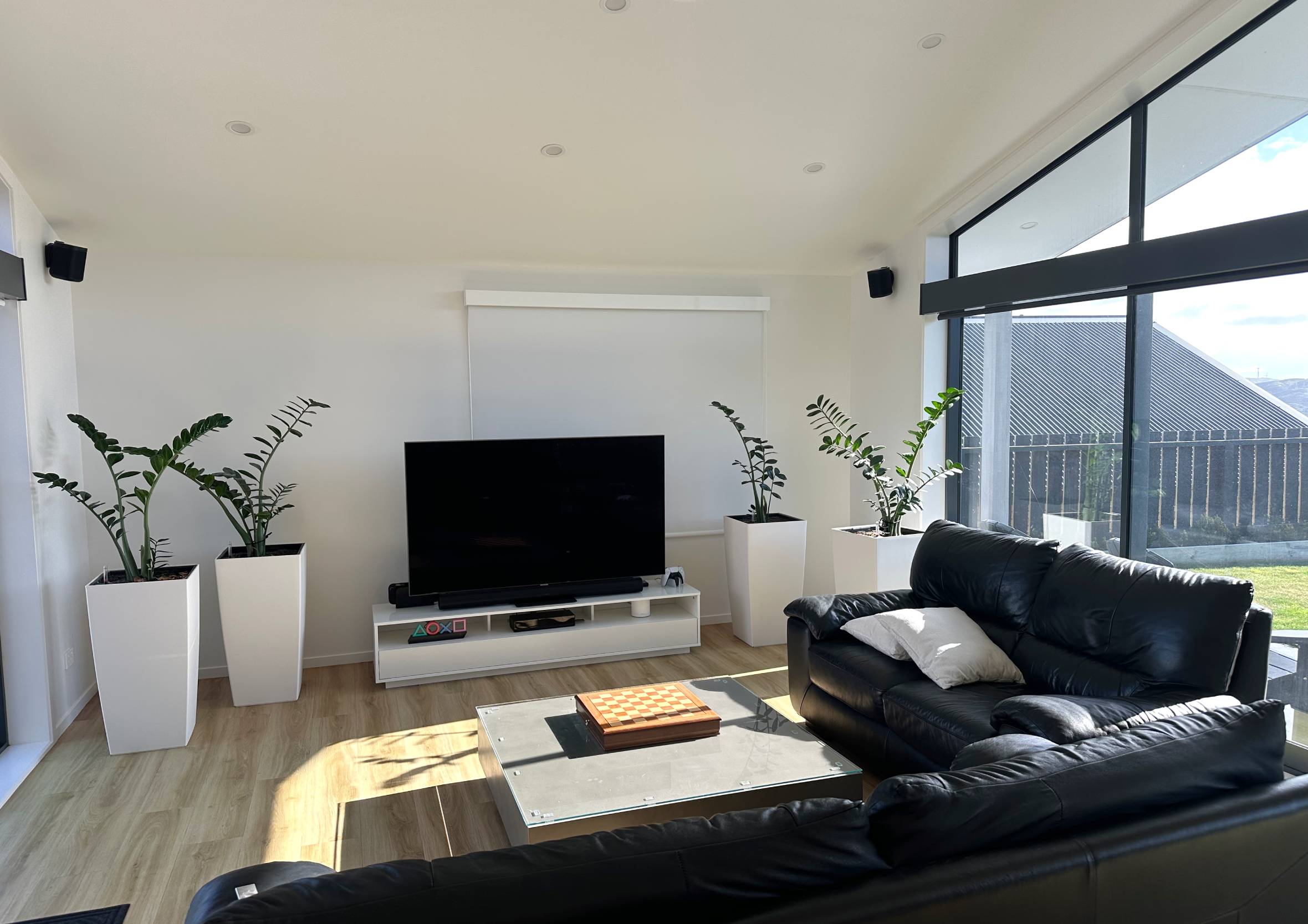 Living room wall layout featuring a low white TV console, television screen, pull-down window blinds, and tall white planters.