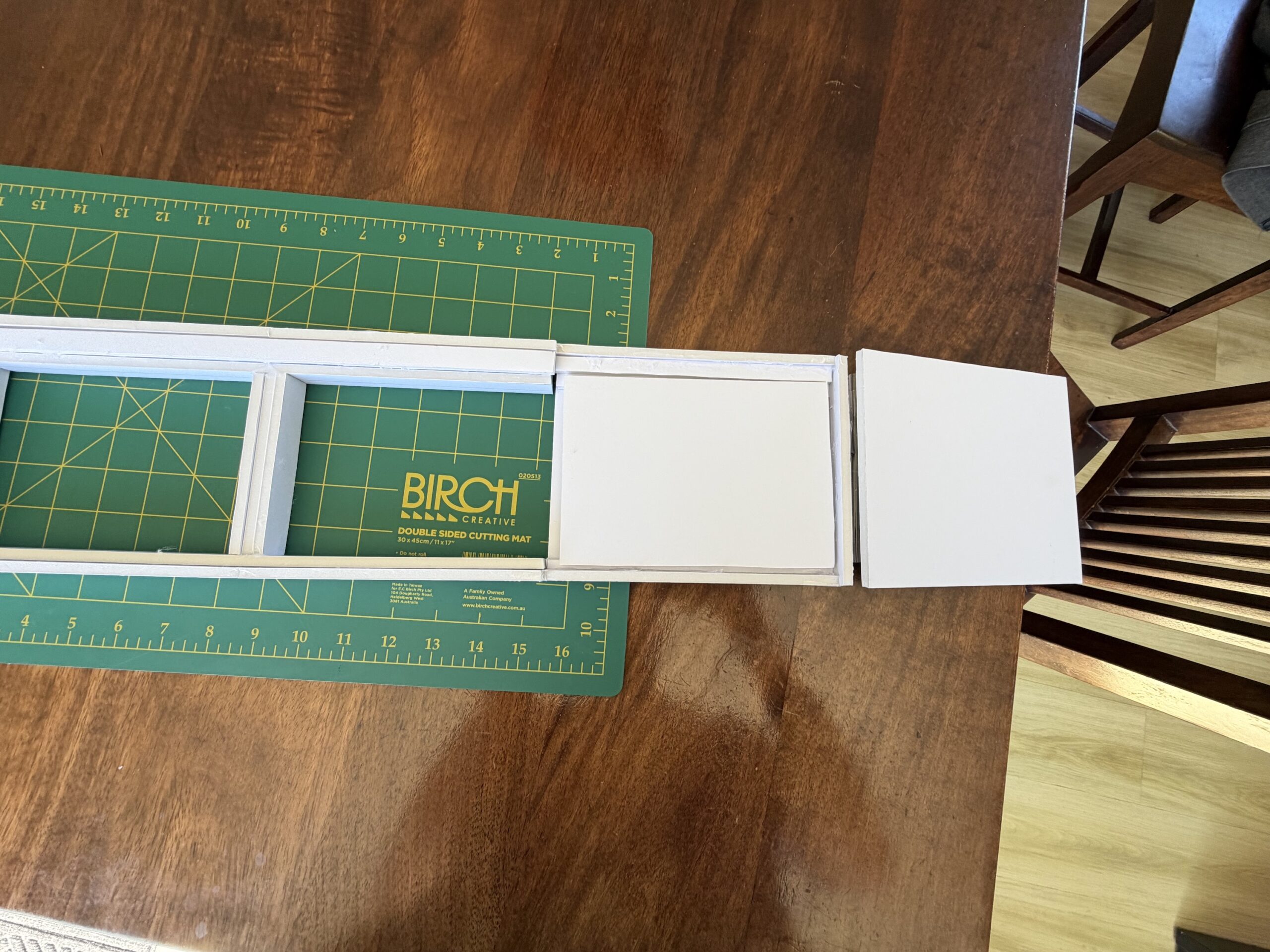 Scale foam-board architectural model of custom Hi-Fi furniture top track system resting on a green Birch cutting mat over a dark timber table, displaying sliding track clearance and hinged lid mechanics.