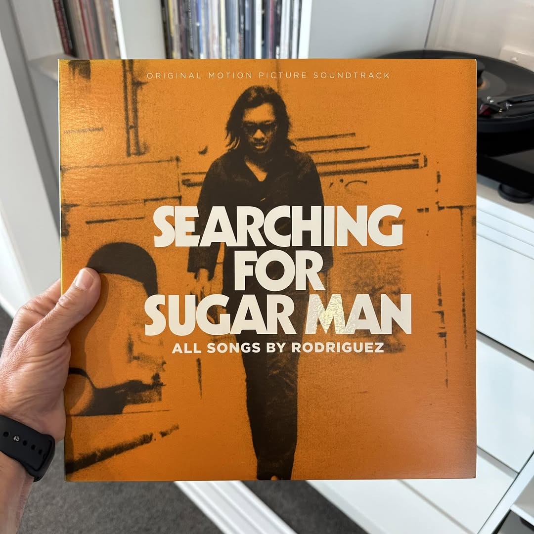 Person holding the Searching for Sugar Man soundtrack vinyl sleeve showcasing a warm orange screen-printed image of Rodriguez walking, paired with micro-copy "All Songs by Rodriguez", capturing vintage documentary visual aesthetics to enrich music feature layouts.