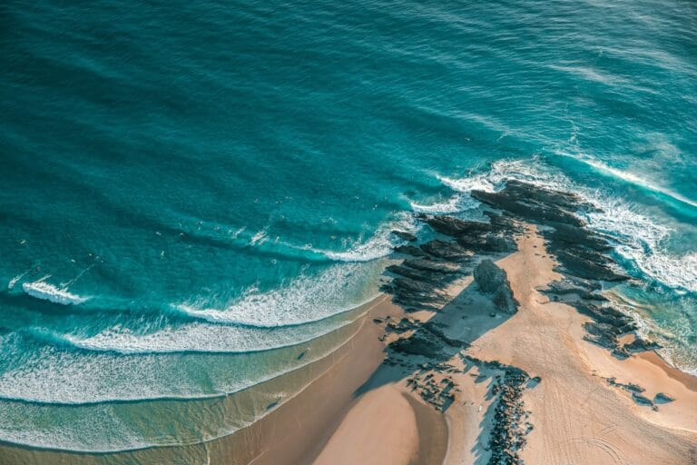 Aerial view of turquoise waves breaking along a beach and rocky headland, surfers floating in the water.