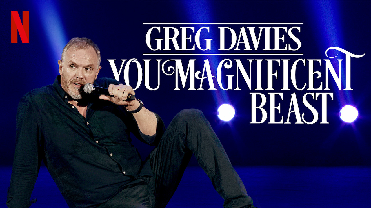 Title card for Greg Davies' stand-up comedy special You Magnificent Beast showing Davies sitting on stage holding a microphone.