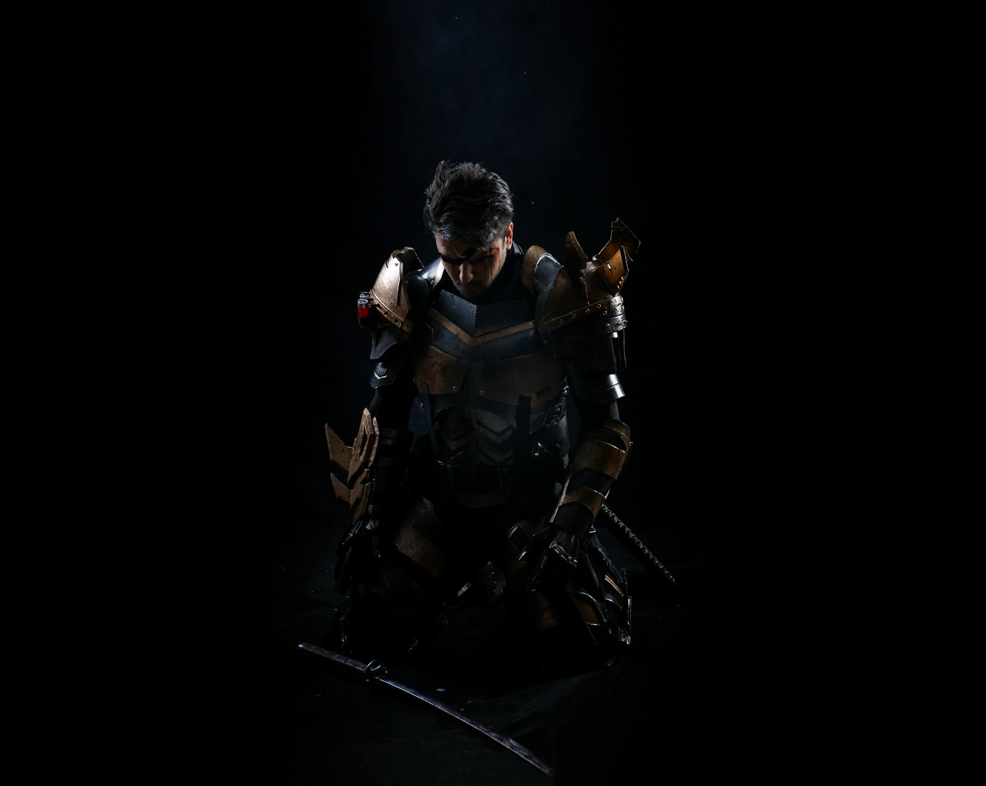 An armoured cosplay warrior kneeling in low light with a katana blade on the ground in front of him.