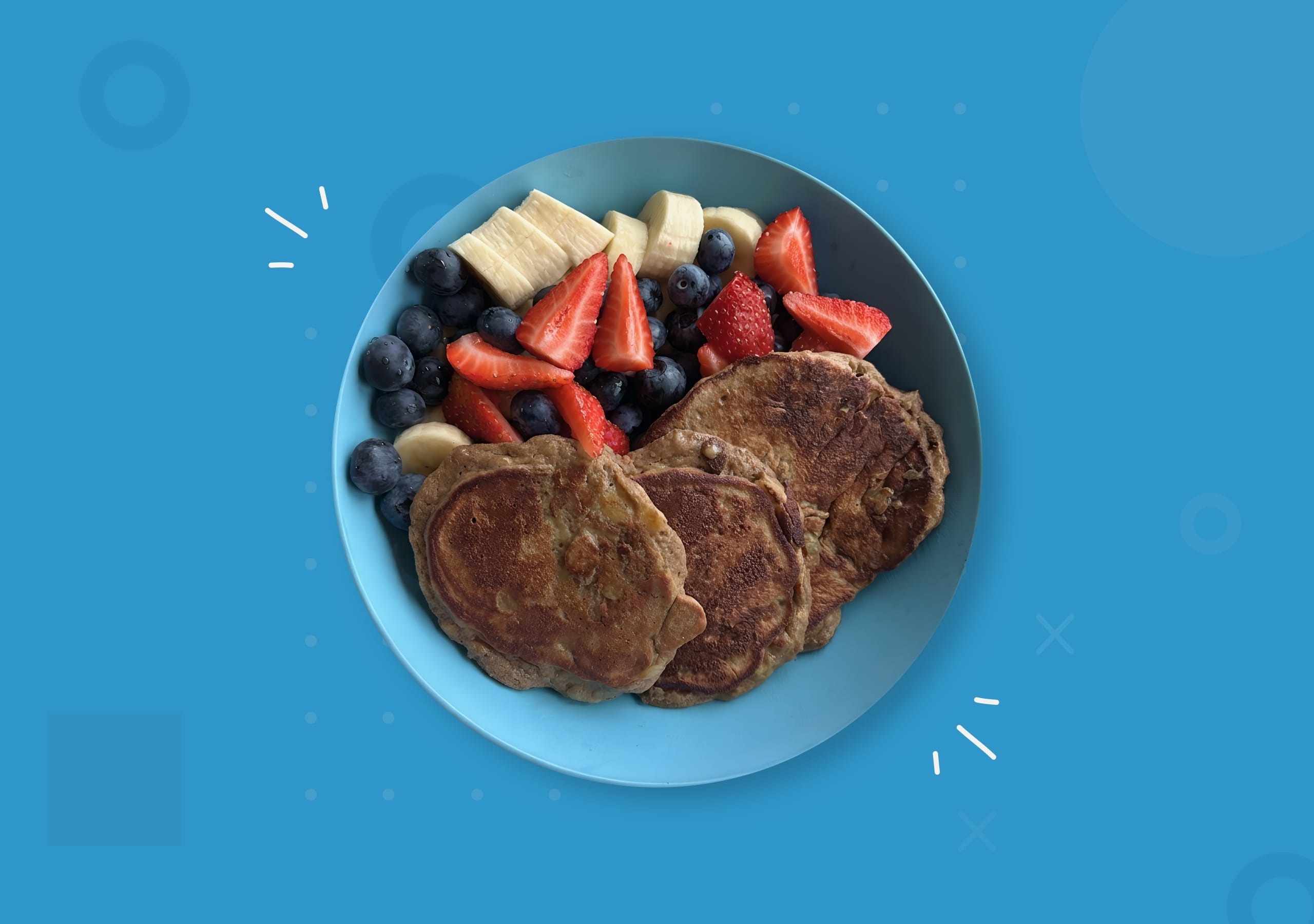 Top-down view of a light blue bowl filled with three fluffy protein pancakes, sliced bananas, fresh blueberries, and chopped strawberries.