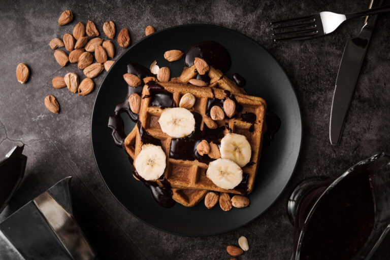Top-down view of golden waffles on a matte black plate, drizzled with dark chocolate sauce and topped with fresh banana slices and whole almonds, set against a dark slate background alongside a fork, knife, and moka pot