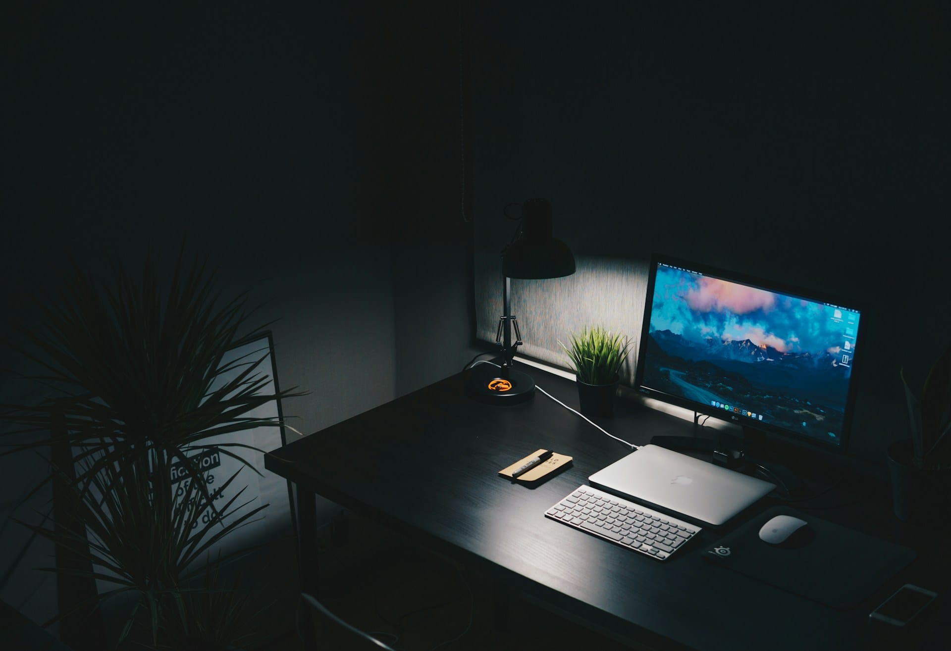 Dark minimalist home office desk setup illuminated by a warm task lamp, featuring a monitor displaying landscape wallpaper, laptop, keyboard, and small potted plant to highlight ergonomic work-life balance workspace essentials.