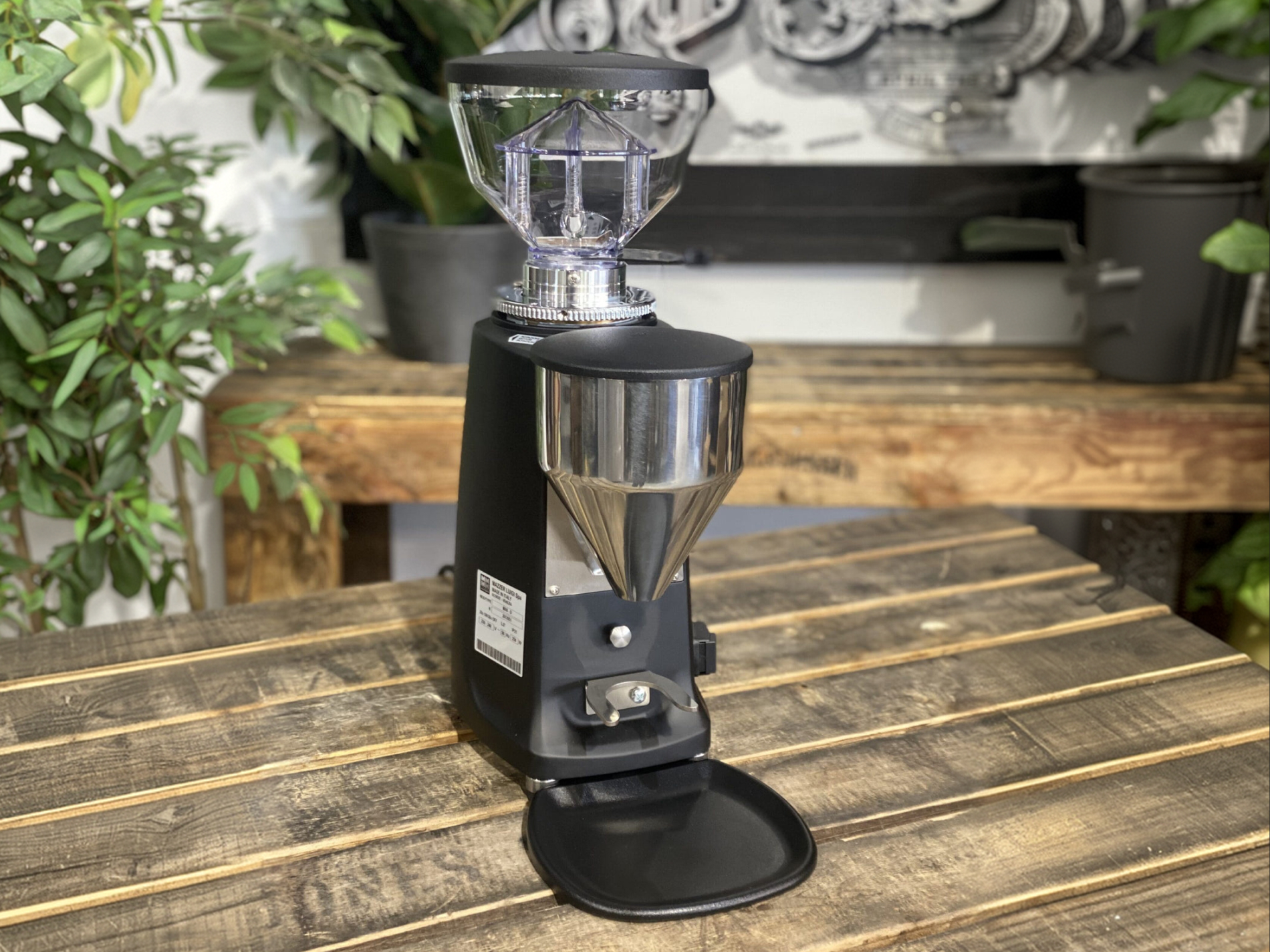 Matte black Mazzer Mini Electronic Model B espresso grinder sitting on a rustic wooden table with a clear bean hopper and stainless steel funnel.