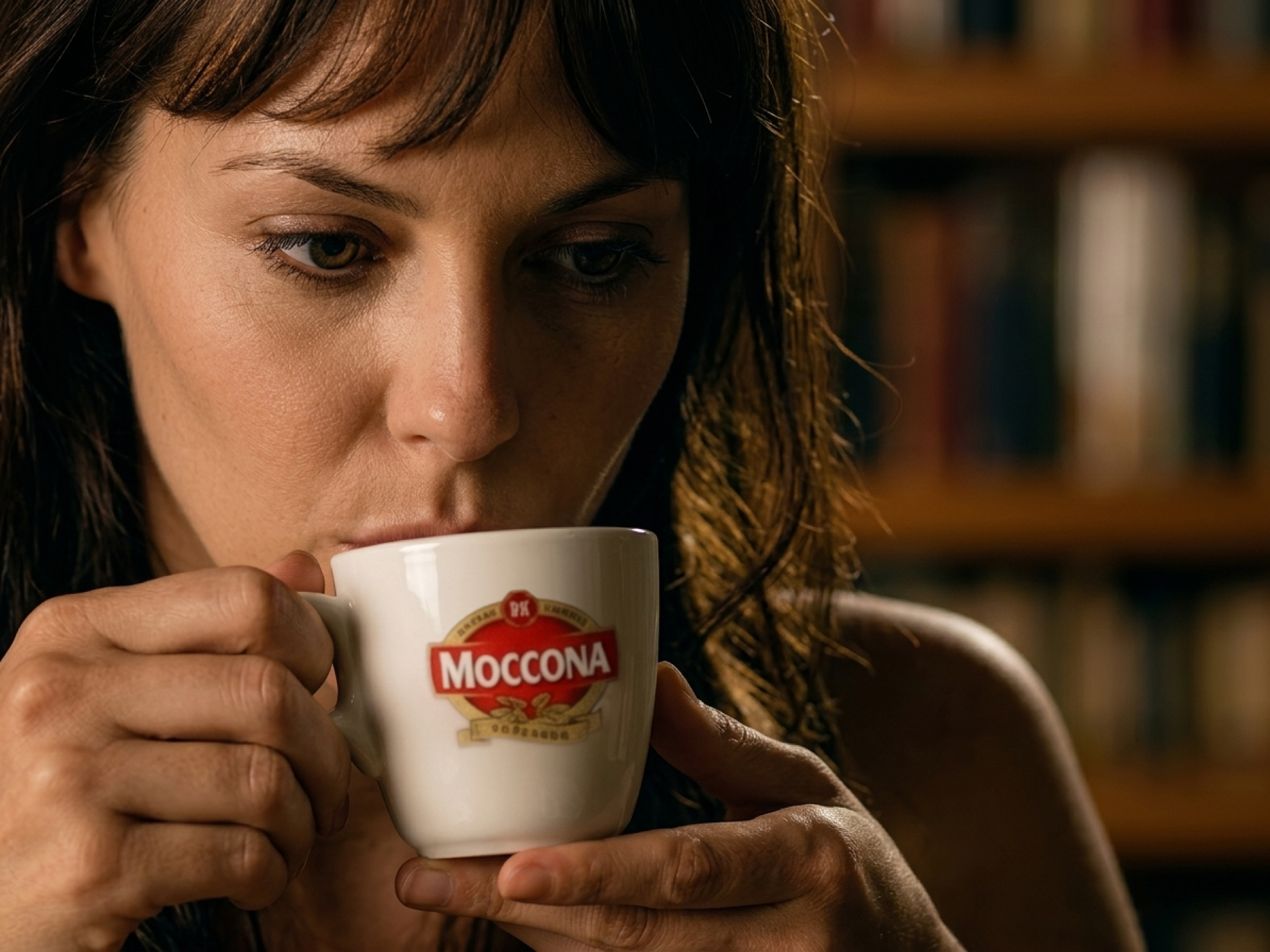A woman drinking coffee from a Moccona-branded cup in a dimly lit room.