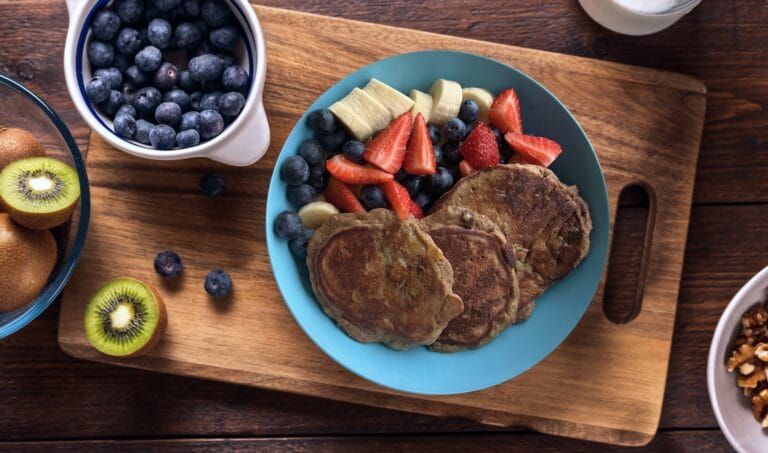 A plate of homemade pancakes topped with sliced strawberries, bananas, and blueberries, served on a wooden board alongside fresh kiwi slices and extra blueberries in a bowl.