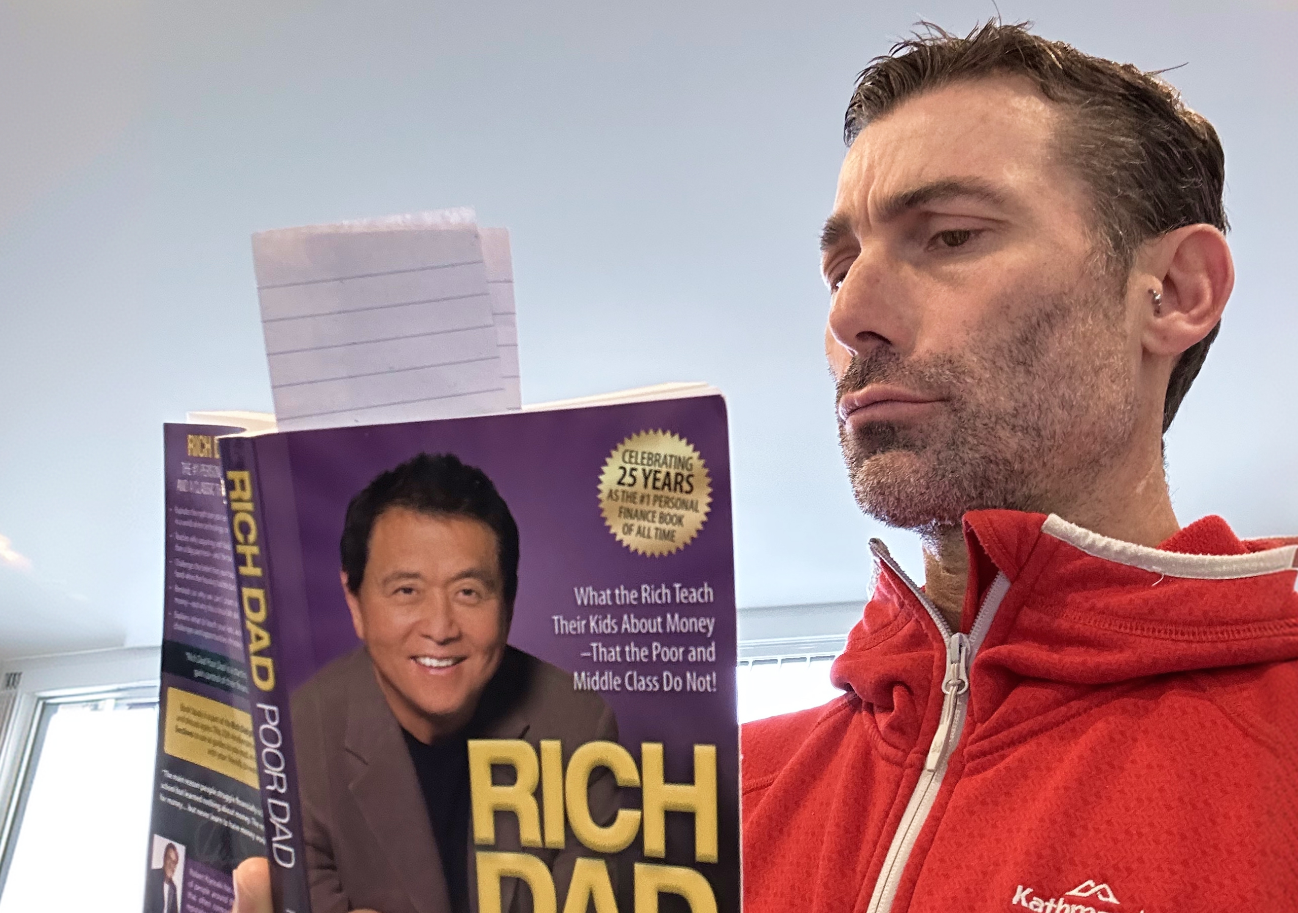 Rich pulling a skeptical face while holding a copy of Rich Dad Poor Dad, a bookmark sticking out the top