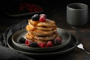 A stack of fluffy pancakes topped with fresh raspberries, blackberries, powdered sugar, and syrup on a dark plate next to a cup of coffee.