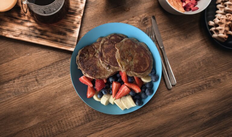 A stack of fluffy protein banana pancakes topped with fresh strawberries, bananas, blueberries on a blue plate next to a cup of coffee.