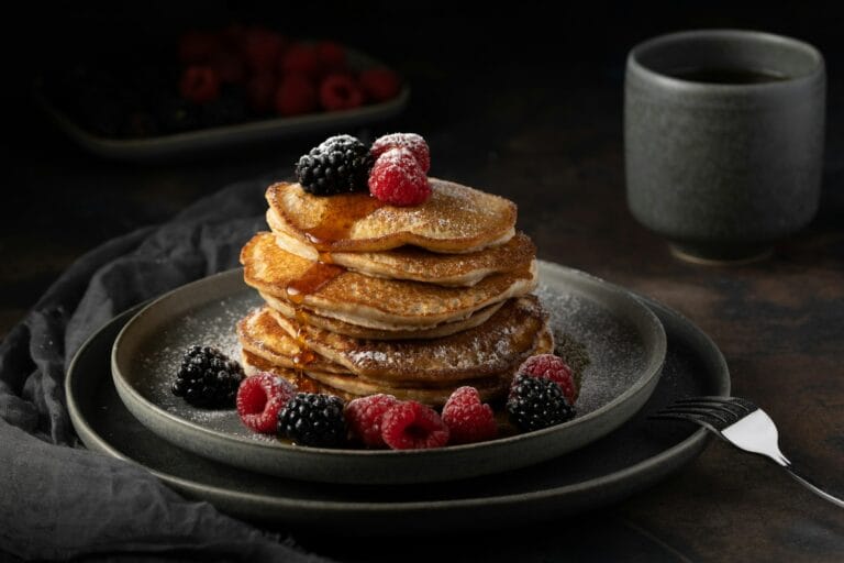 A stack of fluffy pancakes topped with fresh raspberries, blackberries, powdered sugar, and syrup on a dark plate next to a cup of coffee.