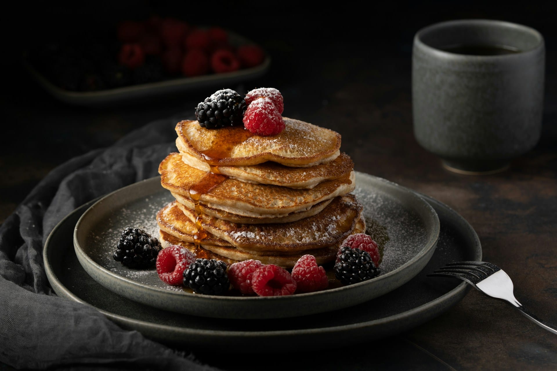 A stack of fluffy pancakes topped with fresh raspberries, blackberries, powdered sugar, and syrup on a dark plate next to a cup of coffee.