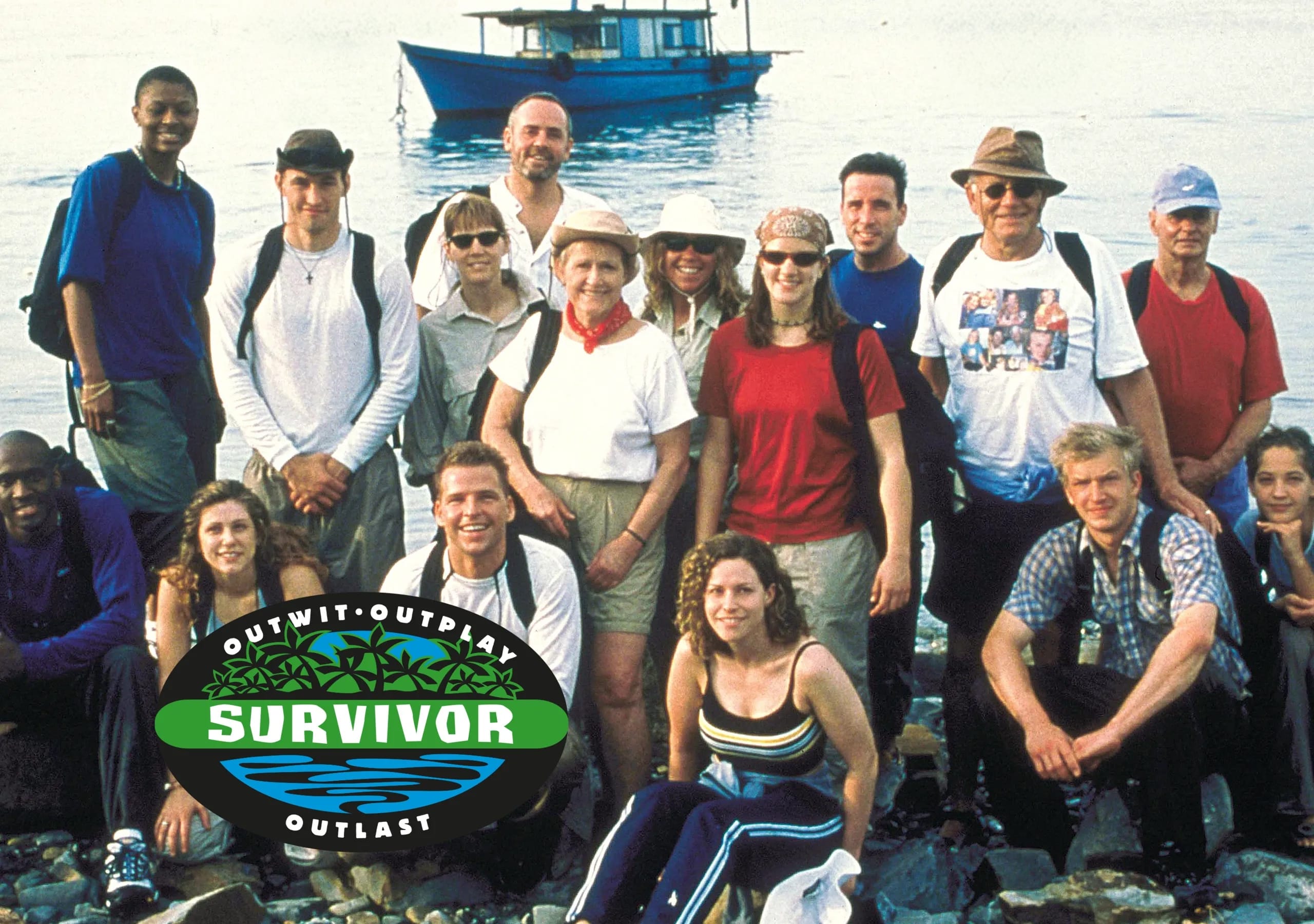 Group photo of all sixteen contestants from Survivor Season 1 Borneo posing together outdoors on a rocky shore in front of the water