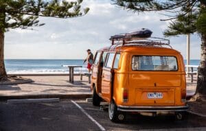 A vintage orange Volkswagen camper van with a roof rack, parked beachside under a cloudy sky.