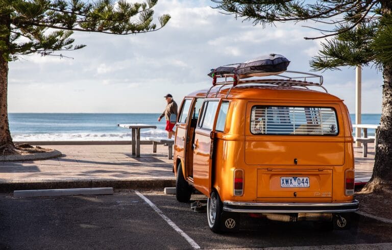 A vintage orange Volkswagen camper van with a roof rack, parked beachside under a cloudy sky.